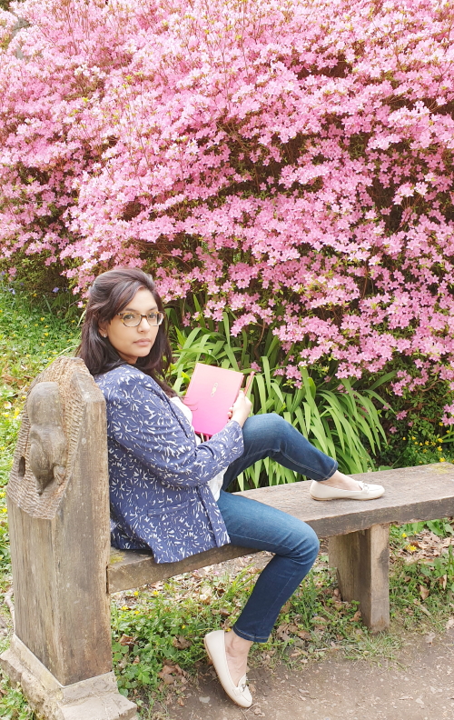 Mona, the co-owner of Acacia & Pine, sitting on a bench surrounded by pink flowers and holding one of her bullet journals